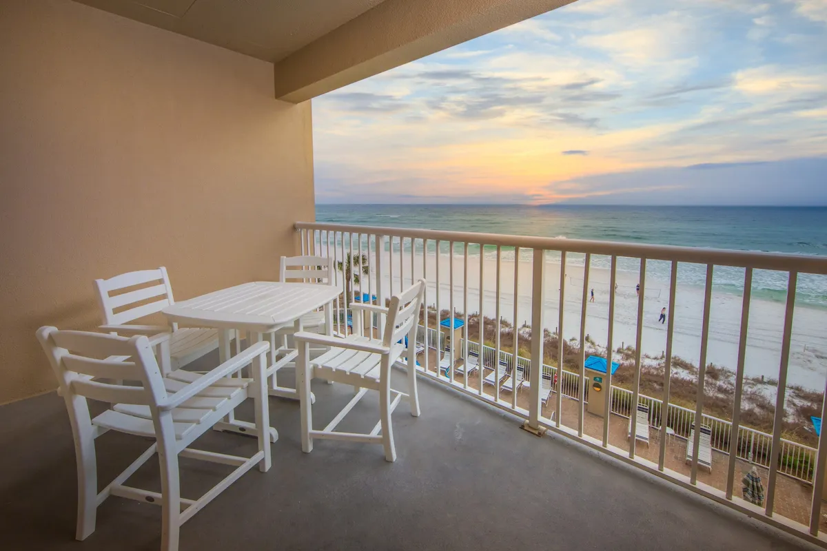 A cozy balcony setup with a white patio furniture set overlooks the beach shoreline, bathed in the warm glow of sunset. The scene suggests relaxation after an active day, with opportunities to dine, unwind with friends, or simply enjoy the peaceful ocean view.
