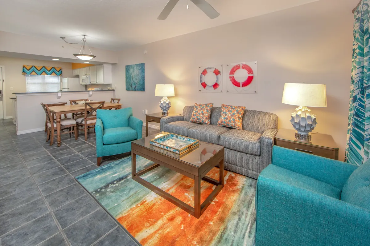 A bright, open-plan interior showcases a striped gray sofa with colorful pillows, two teal armchairs, a vibrant rug, and a wooden dining table with six chairs. The kitchen area is subtly included, emphasizing the space’s functionality for preparing and enjoying meals together.
