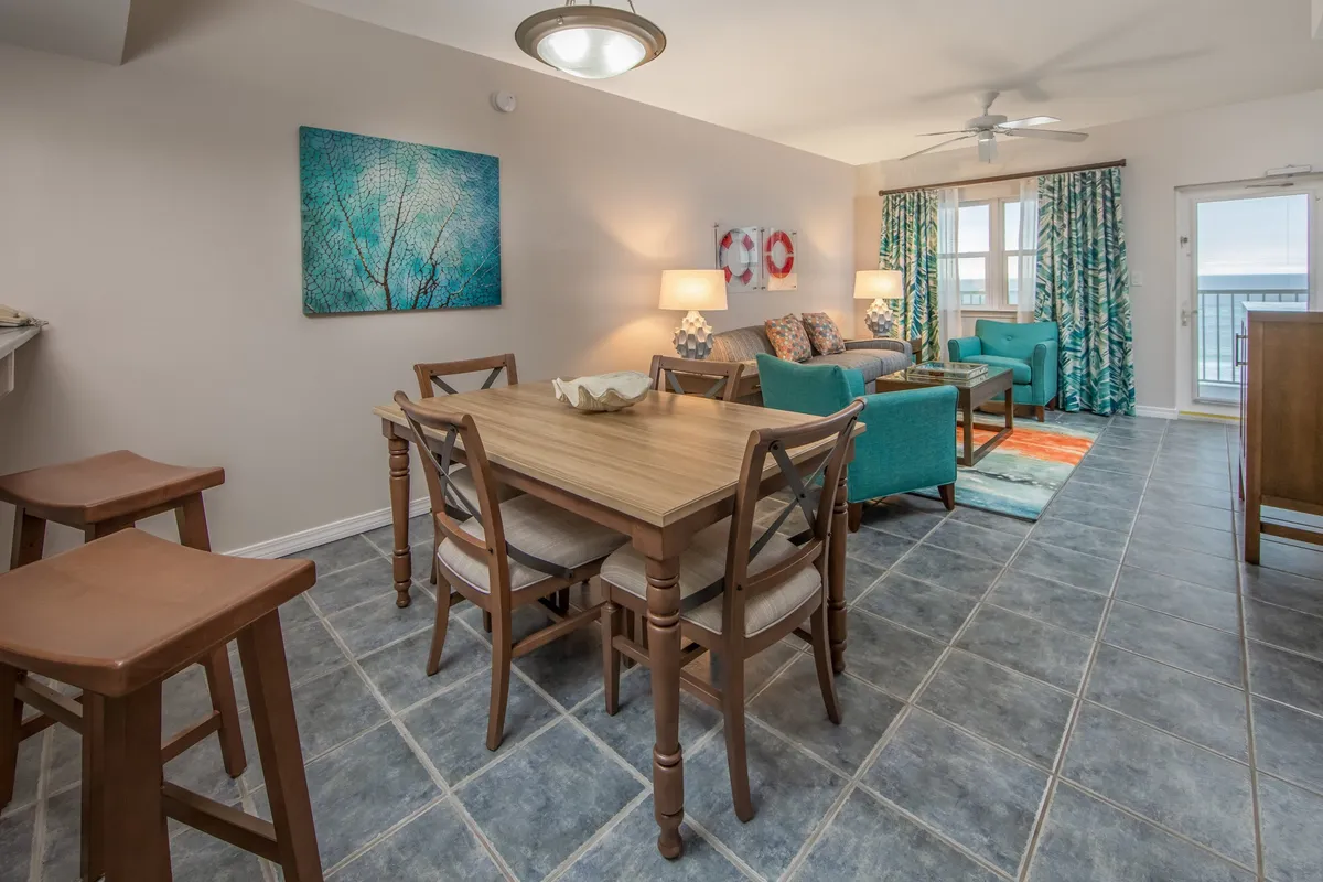 A welcoming dining and living area with a wooden dining table, turquoise armchairs and sofa, colorful wall art, and large windows framing a direct view of the beach. The decor is cheerful and unique, radiating a homey, inviting feel.