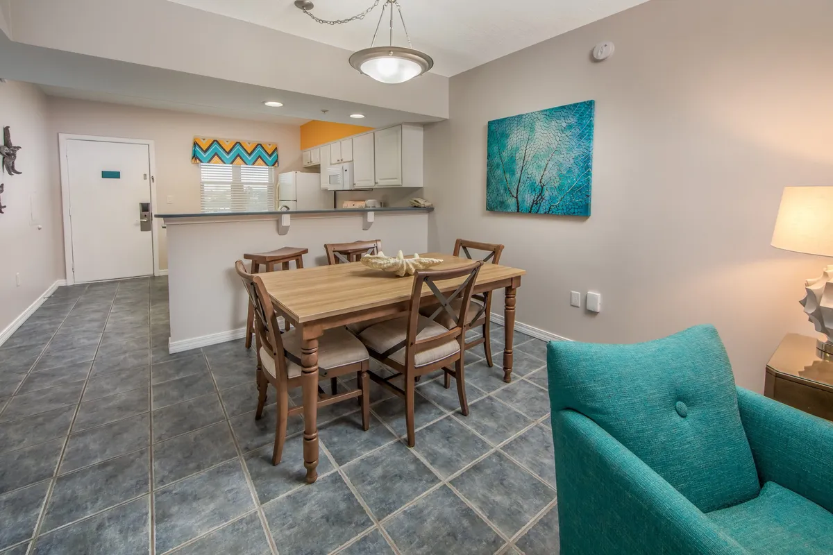This image features a cozy wooden dining table surrounded by chairs, a turquoise armchair for relaxing, and a cheerful kitchen space with a colorful chevron backsplash. Wall art depicting a tree adds a touch of personality. The kitchen is visible in the background, equipped for meal preparation.