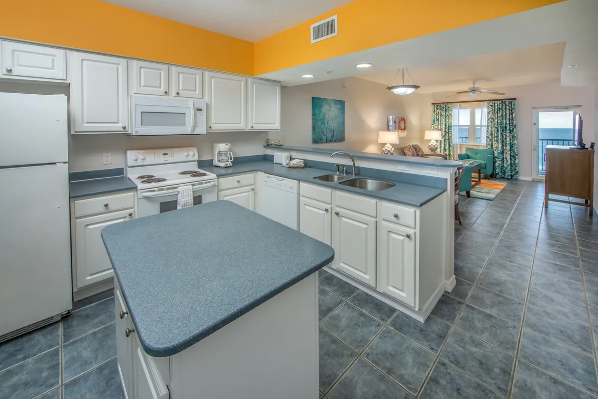 A bright, open-plan living space with a kitchen island, white cabinetry and appliances, and colorful furniture. Large windows and a balcony door offer stunning views of the ocean and beach, while the living area includes a sofa and TV for relaxation.