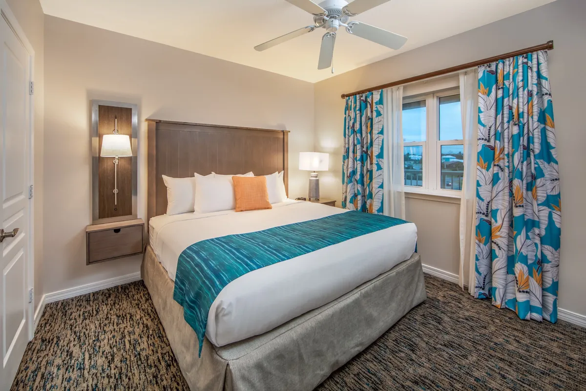 A cozy bedroom featuring a king size bed dressed in blue and white bedding with an orange accent pillow. Natural light streams in through a large window adorned with floral-patterned curtains. The space is finished with a ceiling fan and bedside lamps, creating a restful and inviting ambiance.