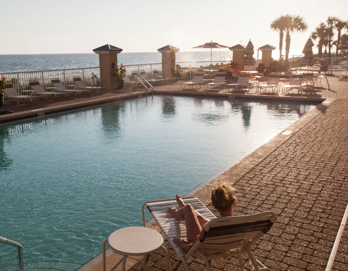 An outdoor swimming pool is the centerpiece, with a person lounging on a pool chair under beach umbrellas. The scene captures the transition from day to dusk, with the ocean horizon and sunset glow in the background. The space is perfect for both relaxing and socializing.
