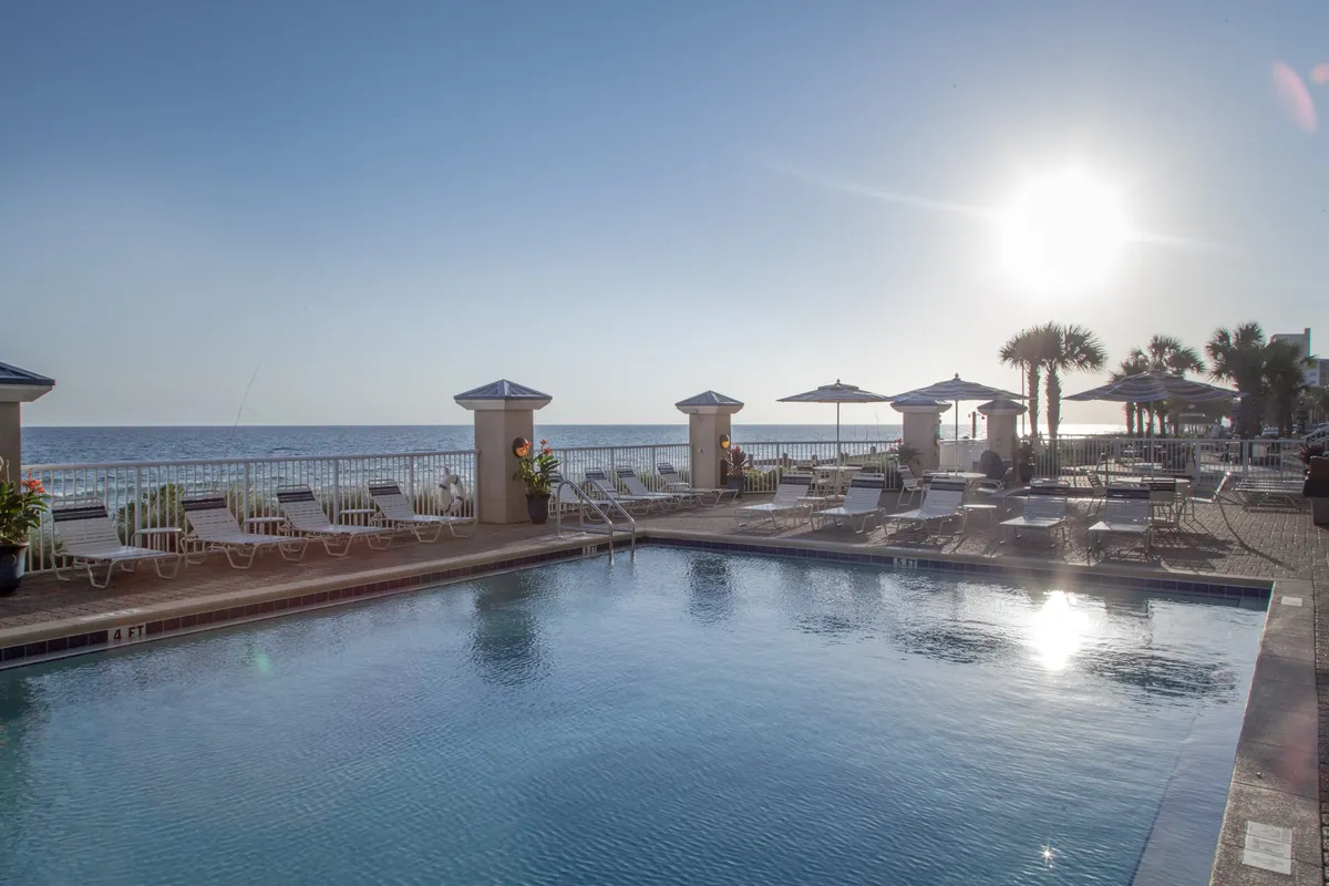 An inviting outdoor pool sits at the center, flanked by plush poolside lounge chairs and umbrella-shaded tables. The sparkling blue water is set against a stunning ocean backdrop, with palm trees and the sandy beach just beyond the pool’s edge. Guests are seen relaxing on lounge chairs, swimming, and socializing around the shaded tables, creating a lively yet peaceful atmosphere.