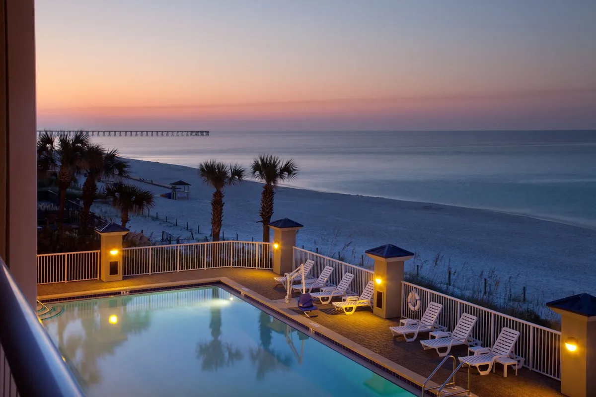An illuminated outdoor pool is surrounded by white lounge chairs and swaying palm trees, with the soft glow of evening light reflecting off the water. In the distance, the beach and ocean horizon are visible, suggesting easy access between poolside relaxation and the sandy shores. The setting evokes a welcoming, family-friendly atmosphere where guests can swim, sunbathe, or gather for evening fun.