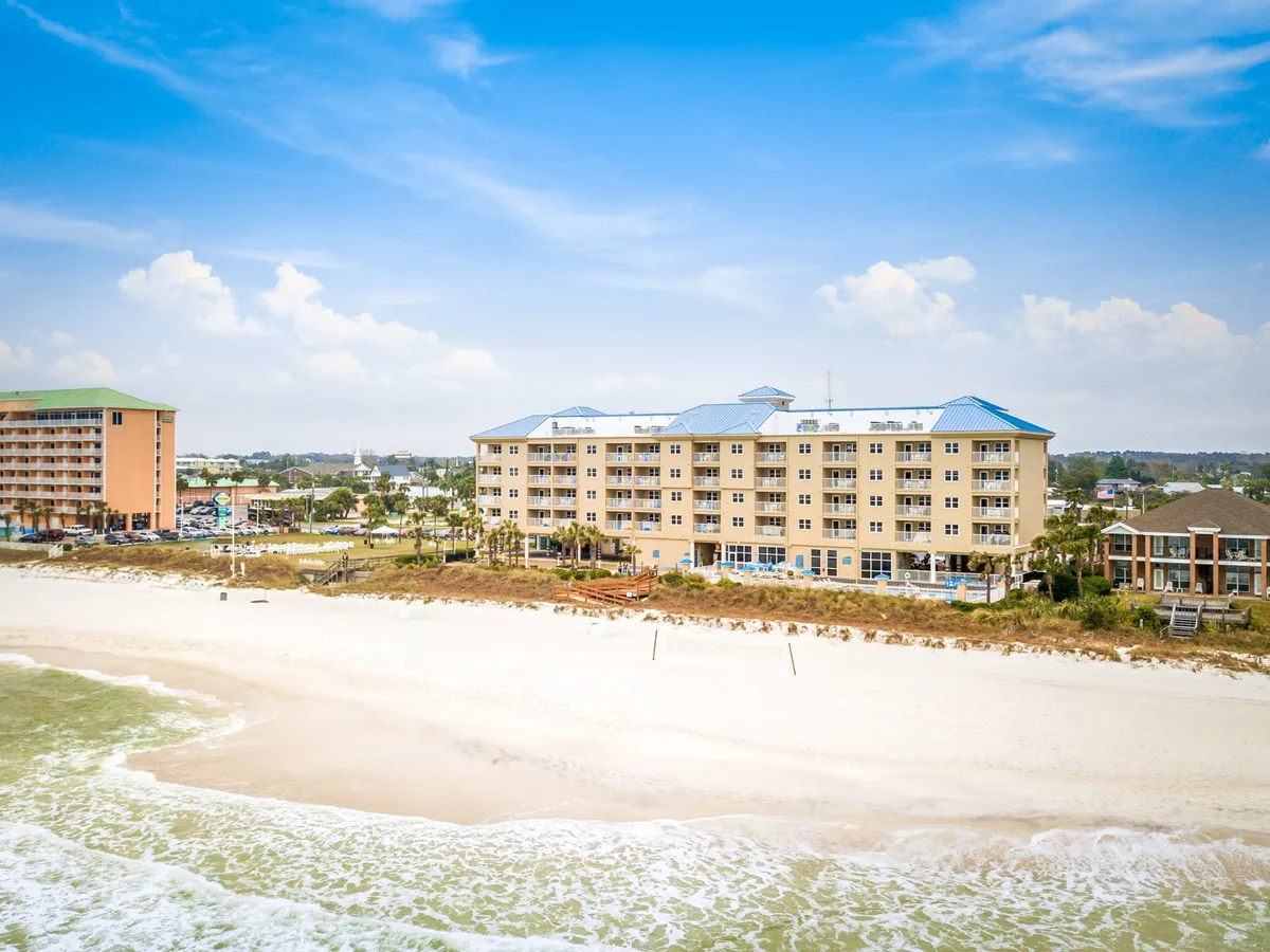 A beachfront villa with a distinctive blue roof sits directly on a wide expanse of sandy white beach, with gentle ocean waves rolling in. Palm trees frame the scene, and adjacent buildings suggest a lively, resort-like environment. The perspective highlights how close the accommodations are to the sand and surf, inviting families to step out and immediately begin their beach adventures.