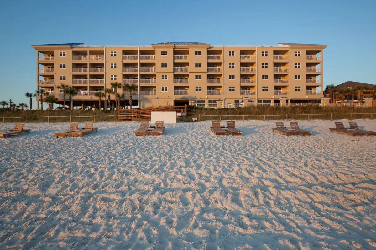 A wide shot of the resort’s modern beachfront villa building, framed by rows of inviting wooden lounge chairs set directly on the sandy beach. In the background, the clear blue sky and the shimmering Gulf create a serene backdrop. The outdoor pool and hot tub are visible, hinting at vibrant social spaces. The scene evokes relaxation and opportunities for both solitude and group gatherings.