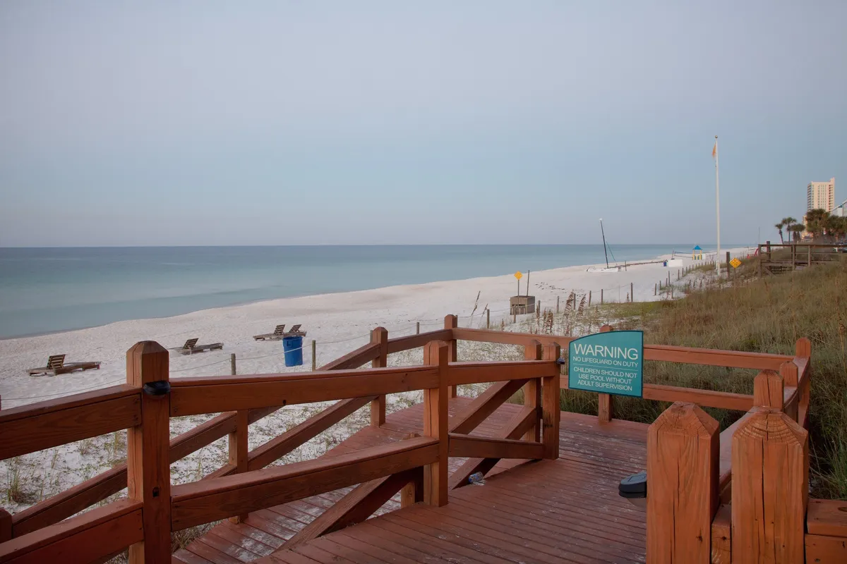 A wooden boardwalk leads directly onto a pristine white sandy beach, meeting the calm blue ocean under a clear sky. Distant beach chairs are spaced along the shoreline, inviting guests to lounge or take a peaceful stroll. The scene exudes tranquility, with no crowds in sight, emphasizing the secluded and relaxing atmosphere accessible just steps from the villa.