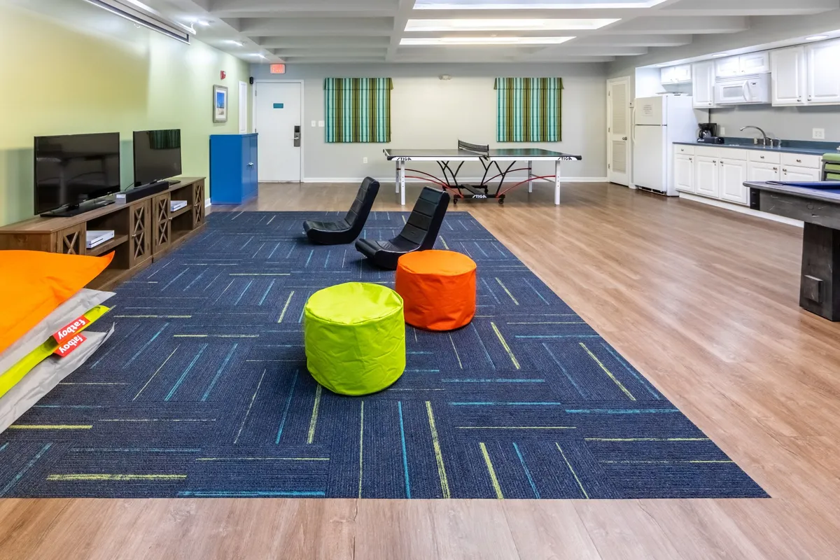 A bright, playful indoor games room featuring a ping pong table at its center, surrounded by comfortable floor chairs and colorful poufs. A large flat-screen TV with a console invites guests to play video games or watch movies. The space is open and inviting, designed for friendly competition, relaxation, and group fun.