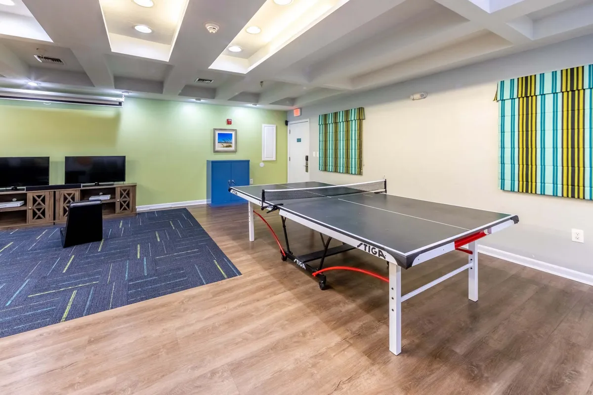 An inviting interior recreation space featuring a ping pong table at the center, two large flat-screen TVs on the wall, blue cabinetry, and playful striped wall decor. The environment encourages socialization, active play, and relaxation, reflecting the family-friendly and fitness amenities available at the resort.