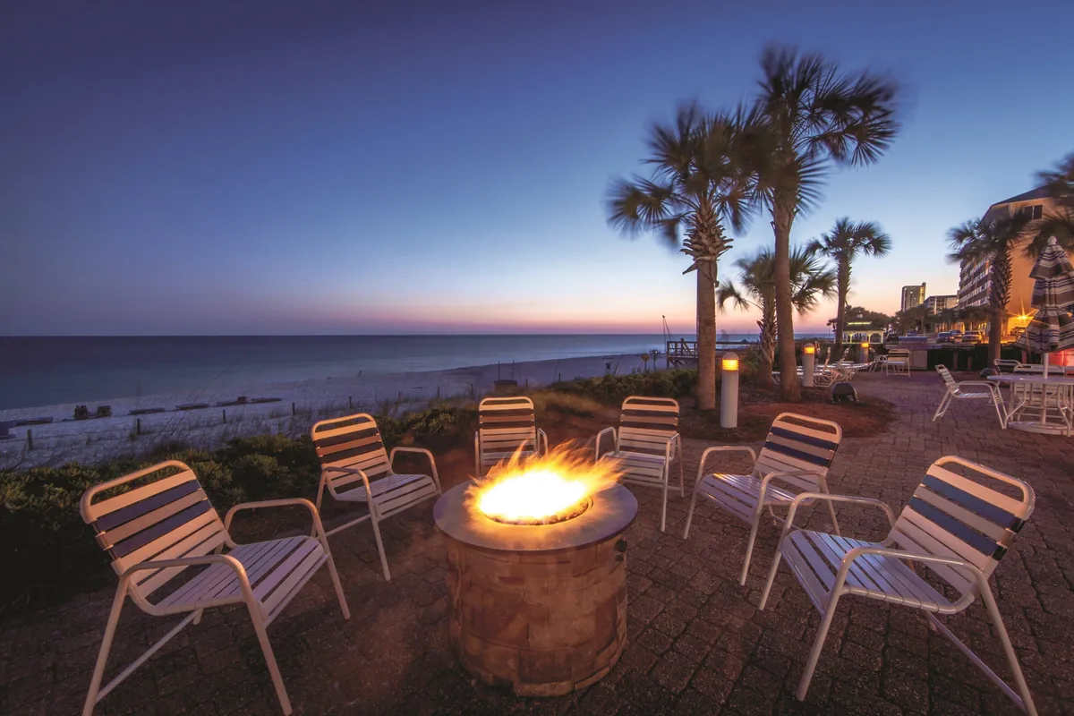 A circular outdoor fire pit glows with burning flames, surrounded by white lounge chairs. Palm trees are silhouetted against a vibrant sunset sky, while the calm ocean and sandy beach create a stunning backdrop. The scene suggests a cozy, inviting atmosphere perfect for unwinding and sharing intimate conversations as evening falls.