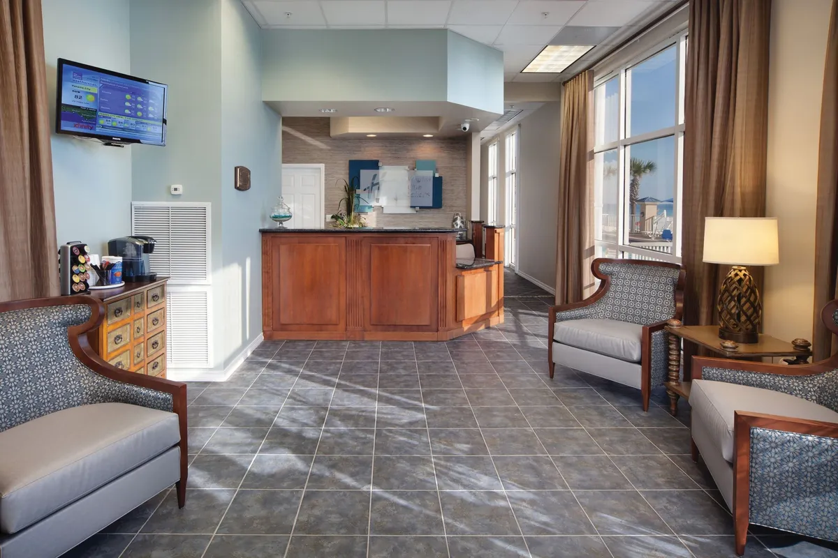 This image depicts the resort's welcoming lobby area, featuring a modern wooden reception desk, plush armchairs for guest comfort, large windows with natural light filtering through curtains, and a wall-mounted TV displaying information. The space exudes calm professionalism and provides a comfortable spot for guests to interact with guest services or relax while waiting.